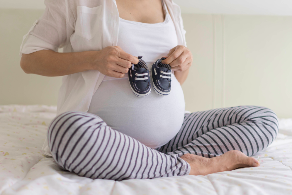 Kobieta w ciąży siedzi na łóżku i trzyma dziecięce buciki